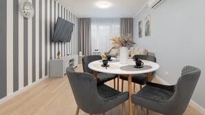 Ein modernes Wohnzimmer mit einem runden Esstisch, grau gepolsterten Sesseln, einem flachbildfernseher an der Wand und einem großen Fenster mit durchsichtigen Vorhängen.