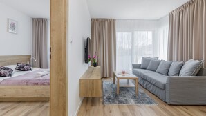 Habitación de hotel moderna con una cama de madera, un sofá gris, una mesita de madera y un televisor fijado a la pared.