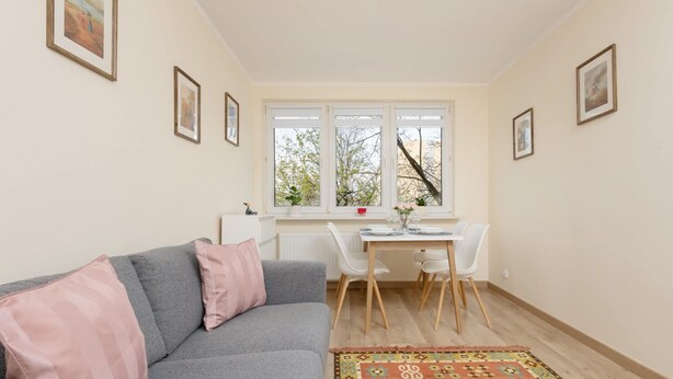 Una sala de estar acogedora con un sofá gris, un tapete estampado y un comedor con una mesa y sillas blancas.