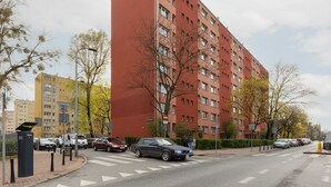 Un edificio residencial de varios pisos con fachada roja, un paso de peatones y coches aparcados a lo largo de la calle.
