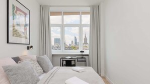 A neatly made bed with pillows, a desk with a lamp, and a window with a city view. - Stylish Apartment in the Center | Air Conditioning (Warszawa)