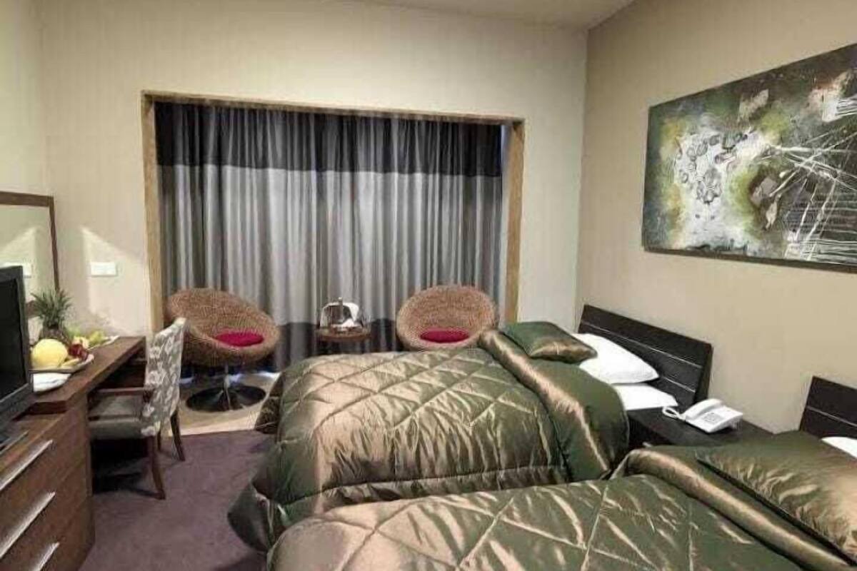 Ein Hotelzimmer mit zwei Betten, einem Schreibtisch mit Computer, einem Stuhl, einem kleinen Tisch mit Lampe und einem Bild an der Wand.