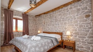 A bedroom with a stone wall, wooden beams, a bed with a grey quilt, two bedside lamps, and a window with curtains. - Lovely home in Donji Vinjani (Donji Vinjani)