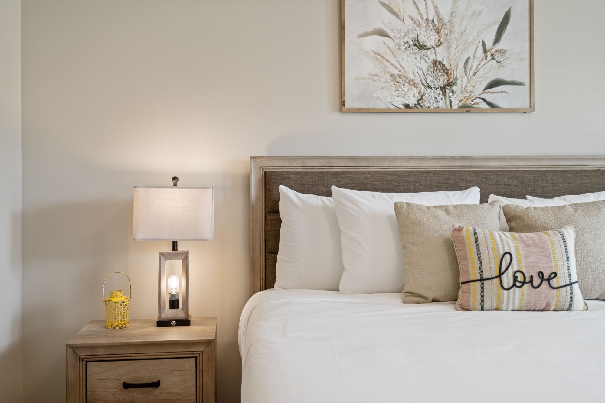 Una cama bien hecha con almohadas blancas y estampadas, una lámpara de noche y una mesita de noche de madera con un farol amarillo.