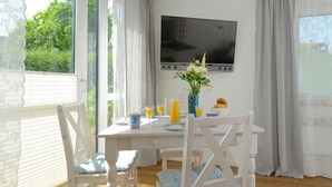 A dining area with a table set for two, a flat-screen TV mounted on the wall, and a window with curtains. - Pretty bungalow with fireplace in Boltenhagen (Ostseebad Boltenhagen)