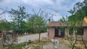 Una casa rústica con techo de tejas, rodeada por un jardín con un sendero empedrado, un pequeño espacio circular de piedra y una valla.