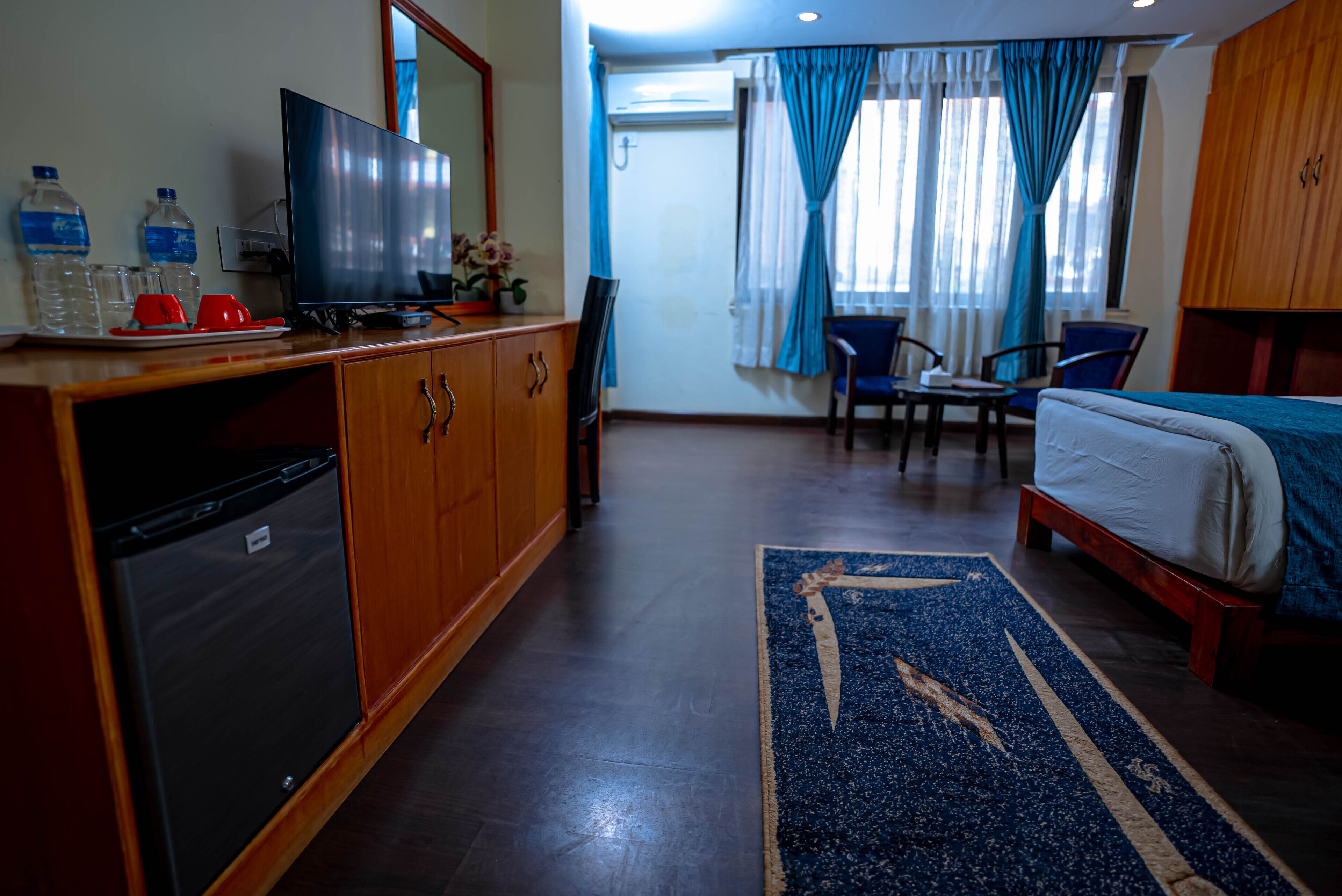 Habitación de hotel con cama, escritorio con silla, televisor, refrigerador y baño visible a través de la ventana.