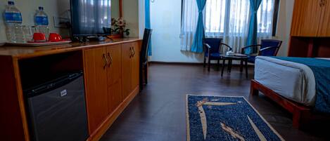 Habitación de hotel con cama, escritorio con silla, televisor, refrigerador y baño visible a través de la ventana.