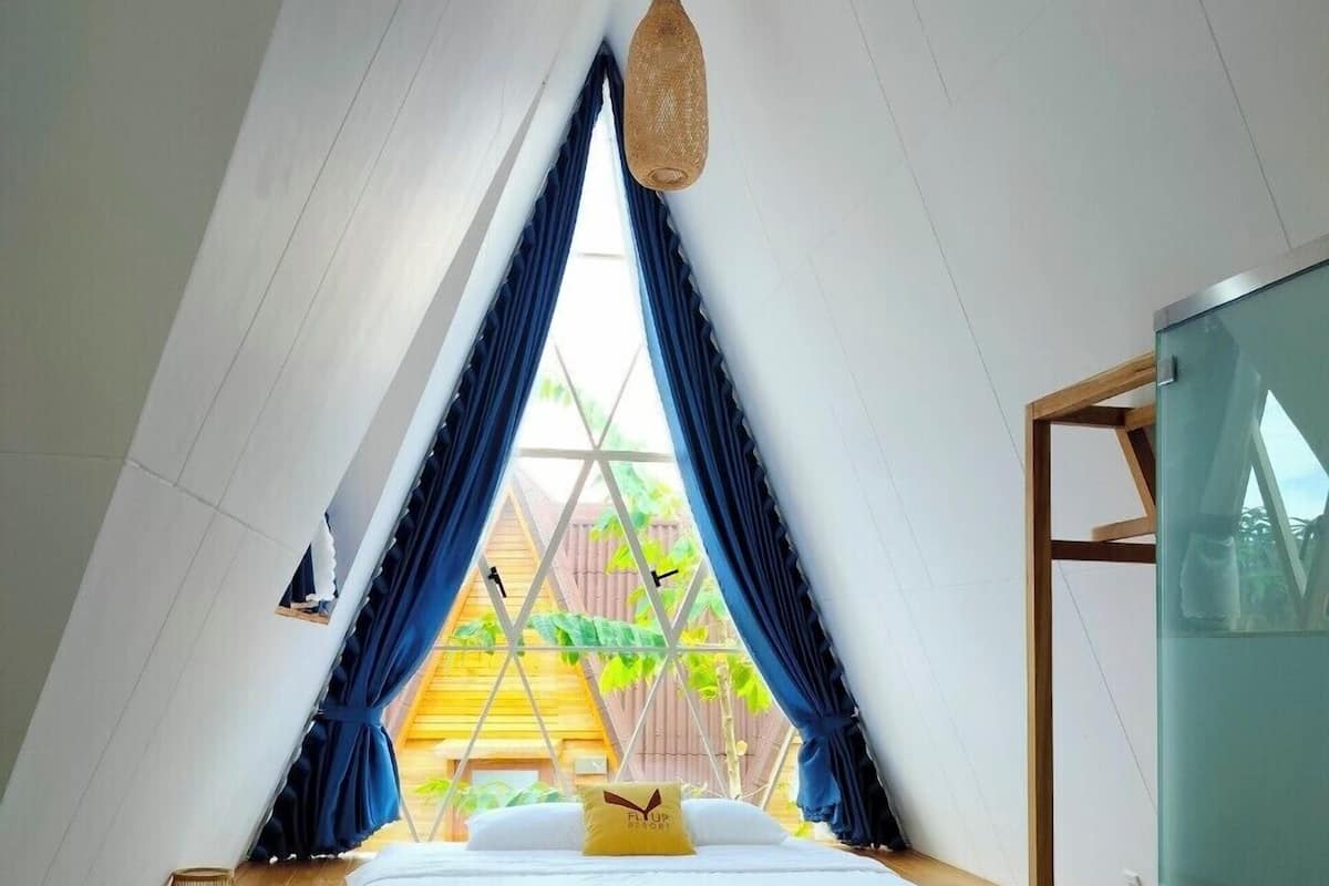 A cozy, triangular attic bedroom with a wooden platform bed, a hanging lamp, and a view of a wooden deck and greenery outside.