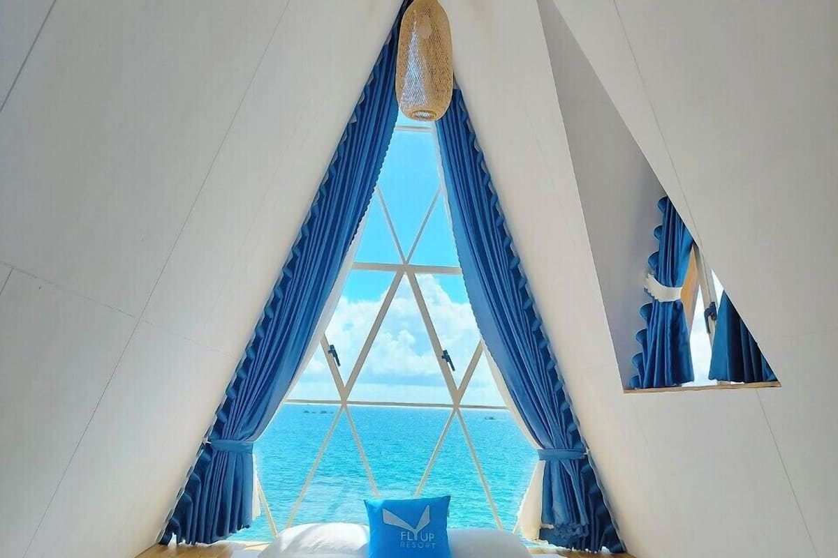 A triangular room with a bed, a hanging light fixture, and a view of the ocean.
