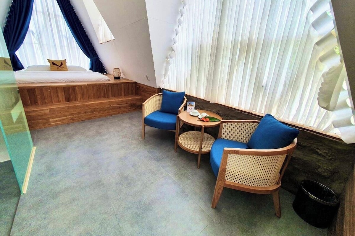 A small, cozy room with a bed, two blue chairs, a small table, and a window with sheer curtains.