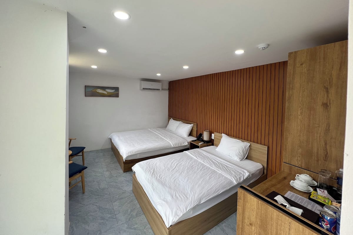 A hotel room with two beds, a wooden wardrobe, a desk with a chair, and a small table with cups and a menu.
