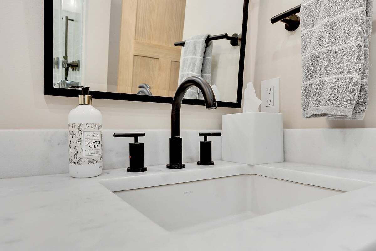 Un baño con un lavabo blanco, grifo negro y una botella de leche de cabra.