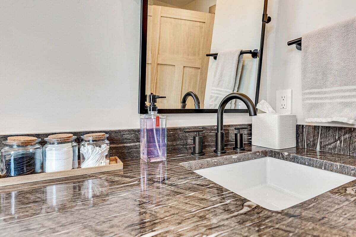 Une salle de bain avec un comptoir en marbre, un lavabo blanc, un miroir, un porte-serviettes et une étagère avec des pots.