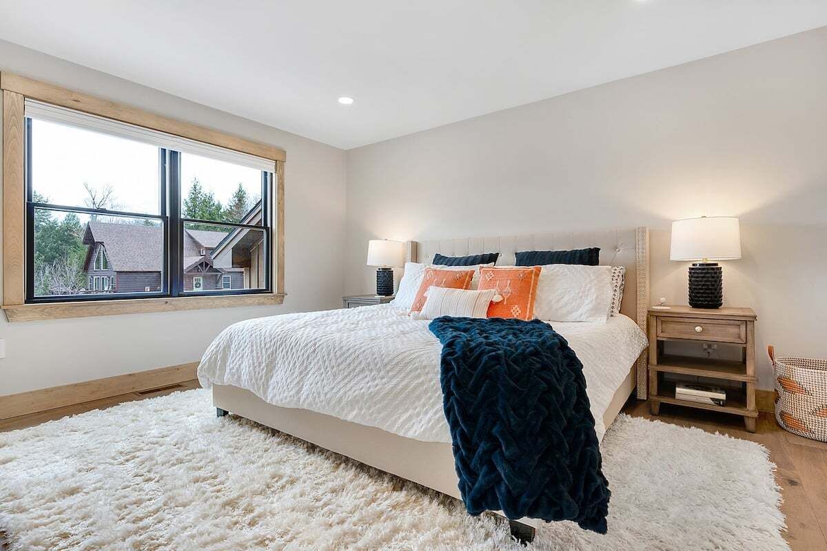 A bedroom with a large window, a bed with multiple pillows, bedside tables with lamps, and a fluffy rug.