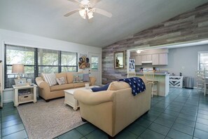 Un salon spacieux avec un canapé beige, une table basse et un ventilateur de plafond. On aperçoit à l’arrière-plan un coin cuisine avec des meubles blancs et une table à manger.
