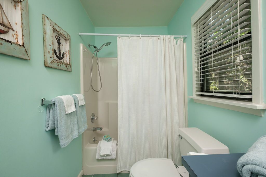 A bathroom with a shower, toilet, and sink. There are towels hanging on a rack and a window with blinds.