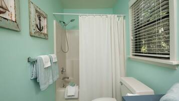 A bathroom with a shower, toilet, and sink. There are towels hanging on a rack and a window with blinds.