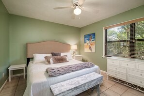 Une chambre à coucher comprenant un lit, des tables de chevet, un banc, un ventilateur de plafond, une fenêtre avec des stores et un tableau accroché au mur.