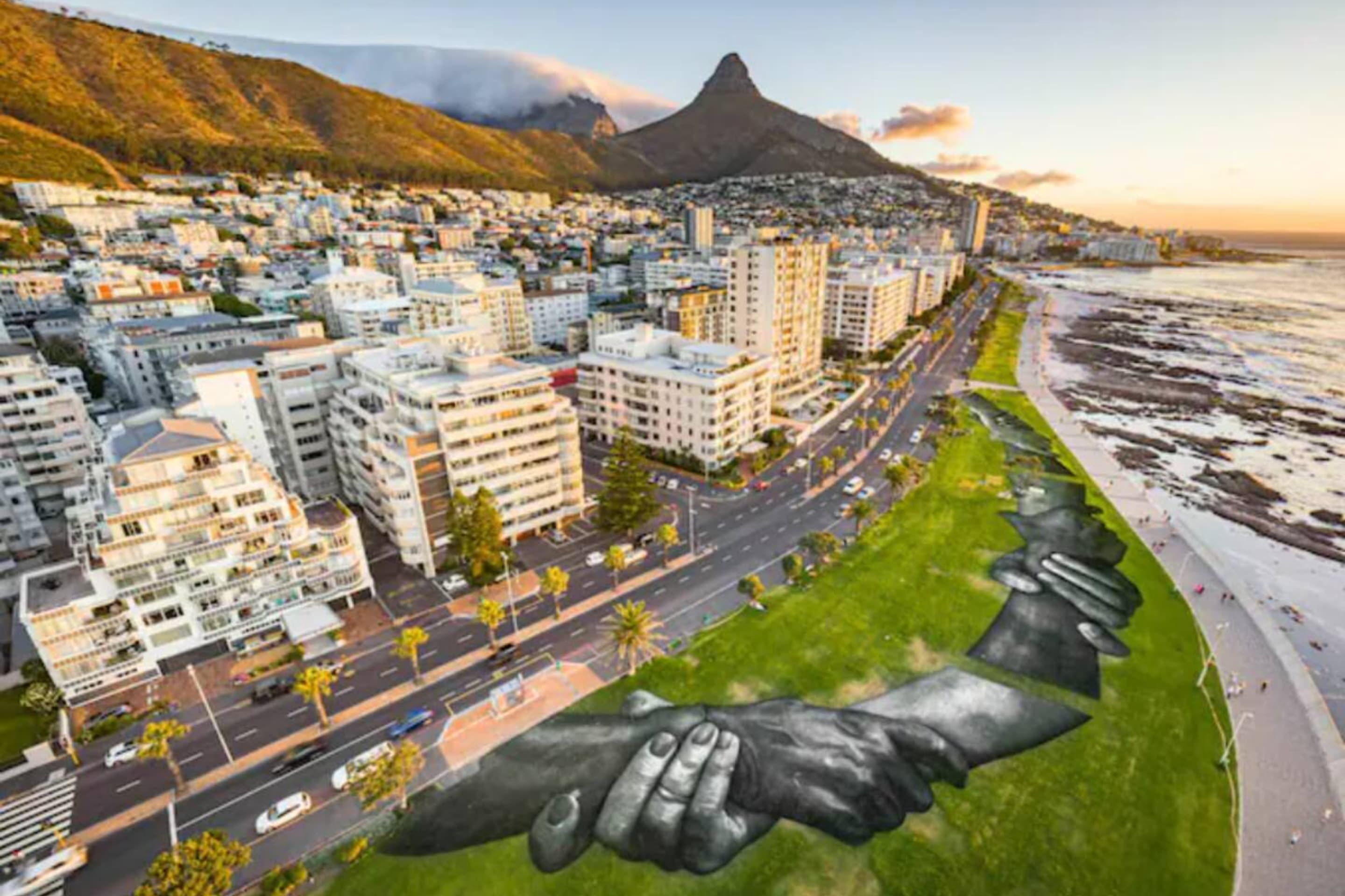 A coastal cityscape with a prominent mountain in the background, a road lined with buildings, and a large sculpture of two hands holding each other.