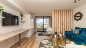 A modern living room with a flat-screen TV, a blue sofa, a wooden accent wall, and a view of the outdoors. - Colourful and Cosy Studio Apartment in Newlands (Cape Town)