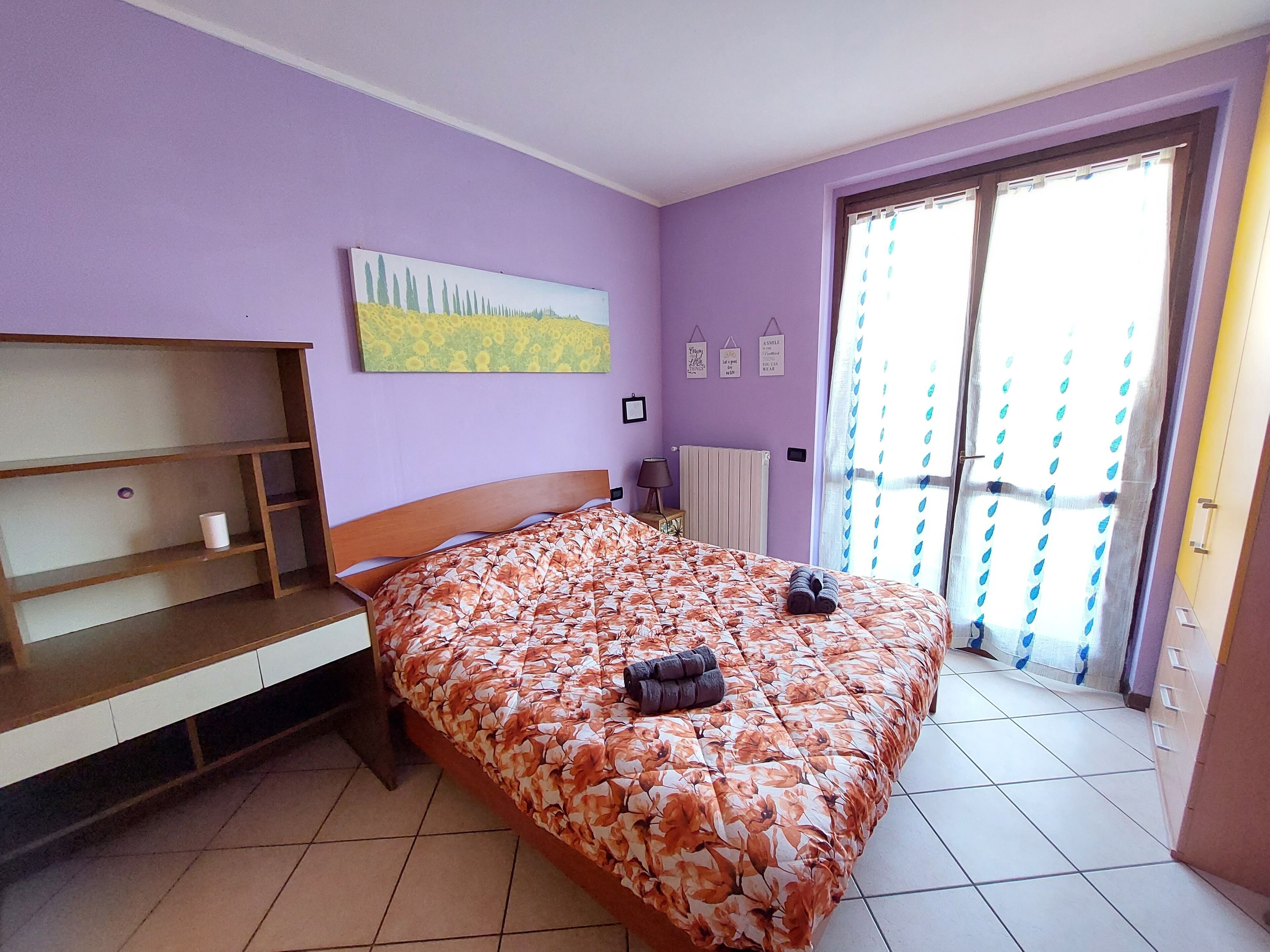 Une chambre à coucher avec un lit, une étagère en bois, un tableau, un radiateur et une fenêtre avec des rideaux.