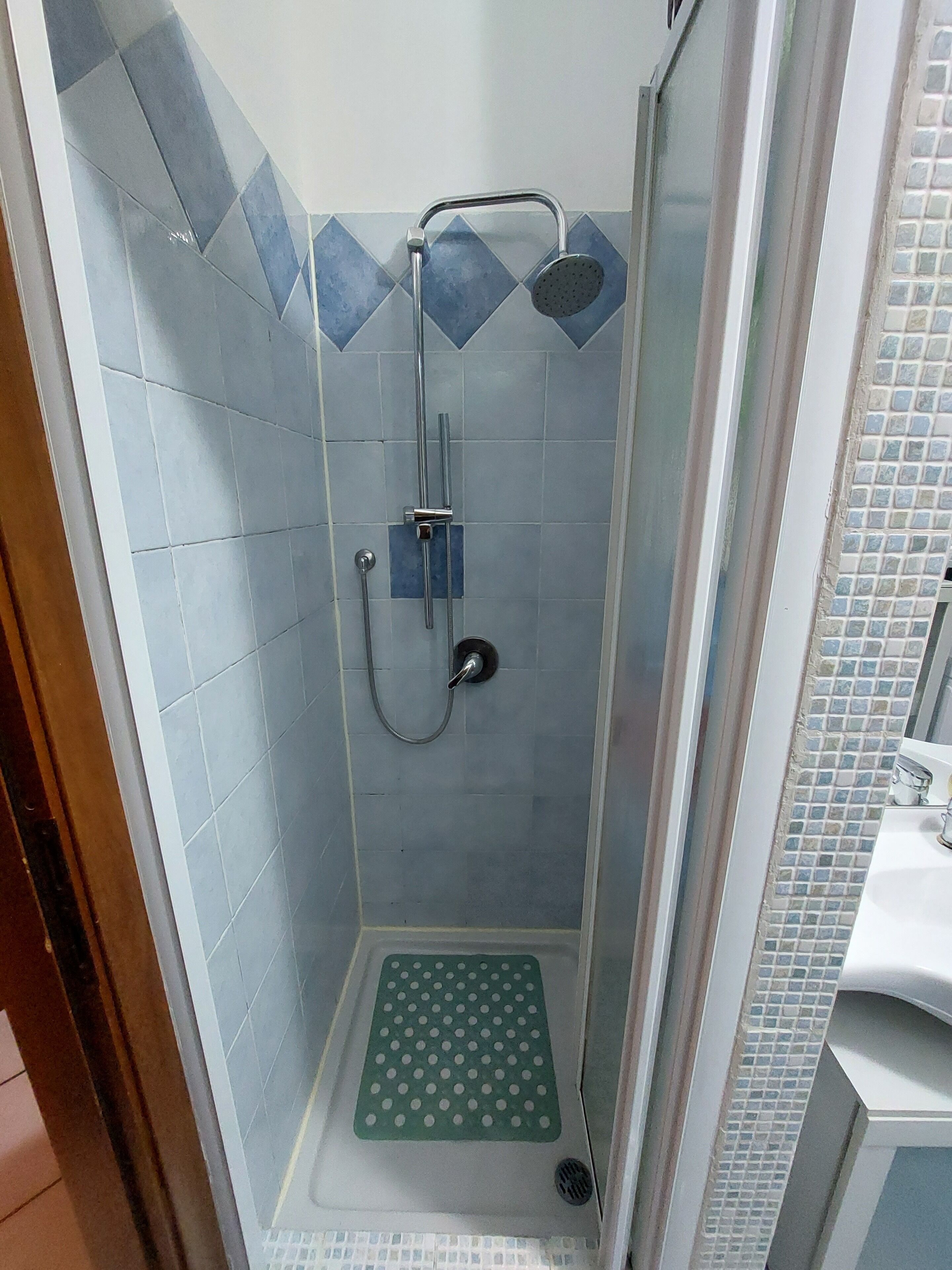 Piccola cabina doccia con tappetino verde, soffione a mano e rubinetto a parete.