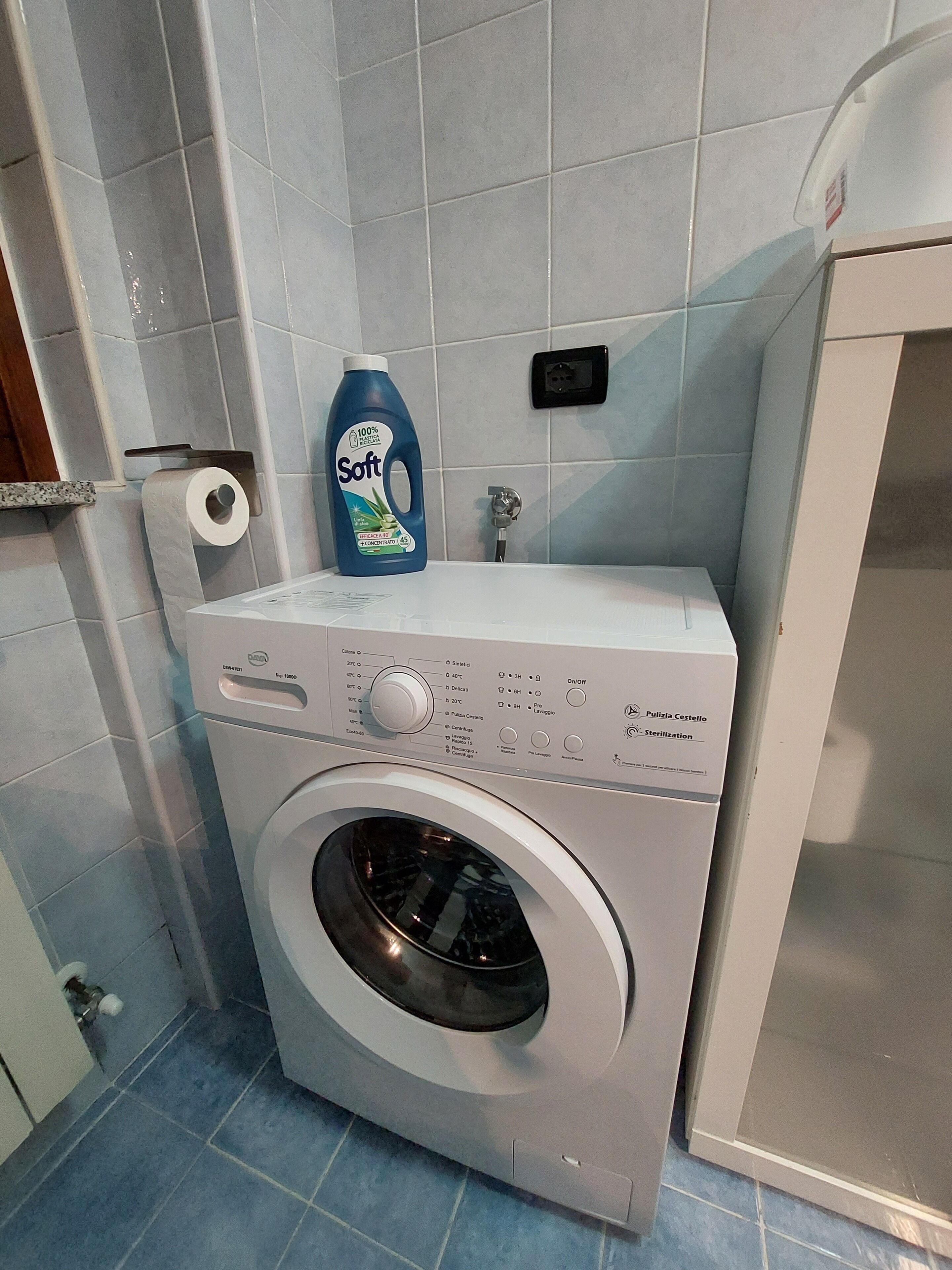 Een wasmachine met een voorlaadklep, een bedieningspaneel, een fles wasmiddel en een papieren handdoekdispenser.