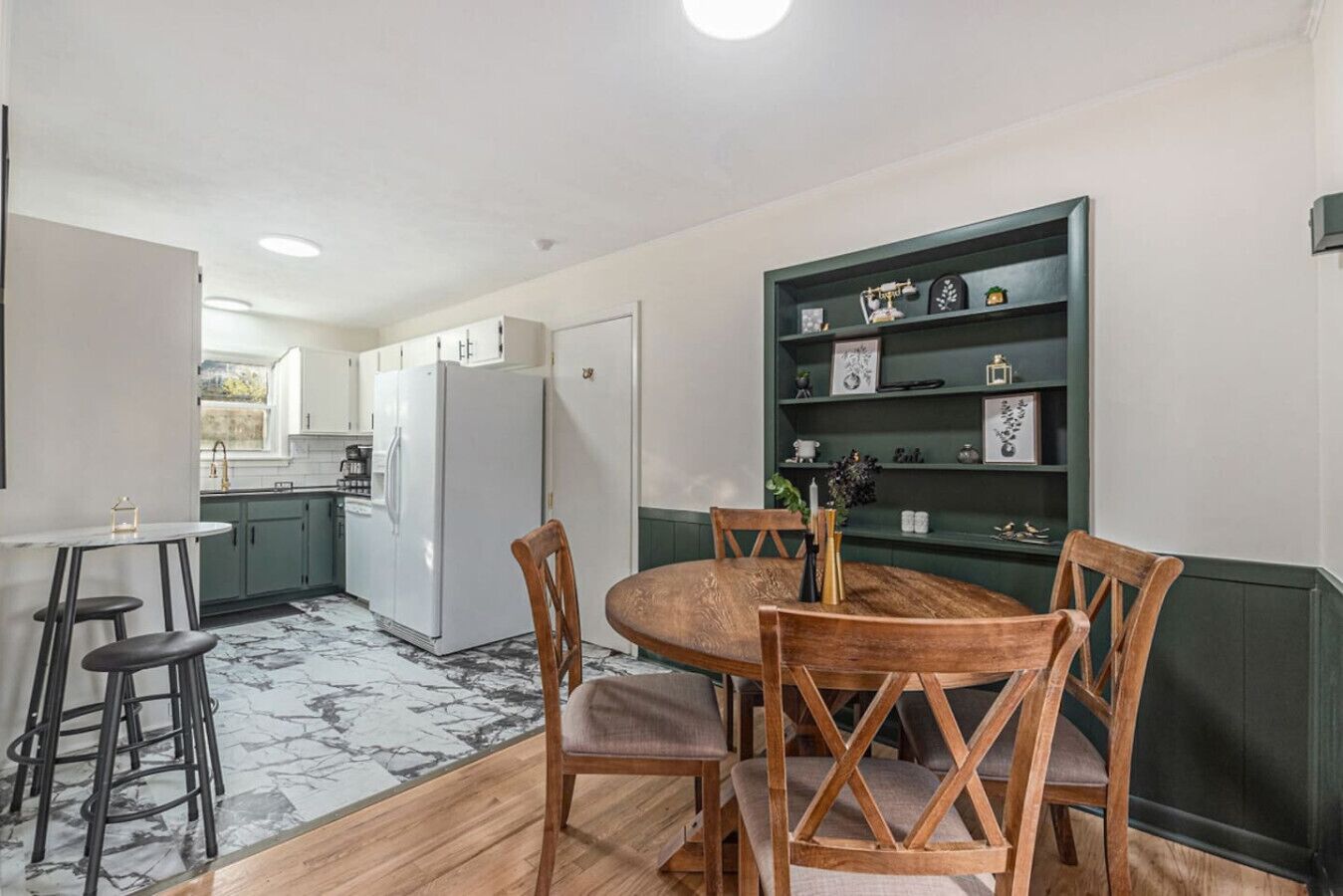 A compact kitchen with white appliances, a dining area with a round table and wooden chairs, and a green bookshelf with decorative items.