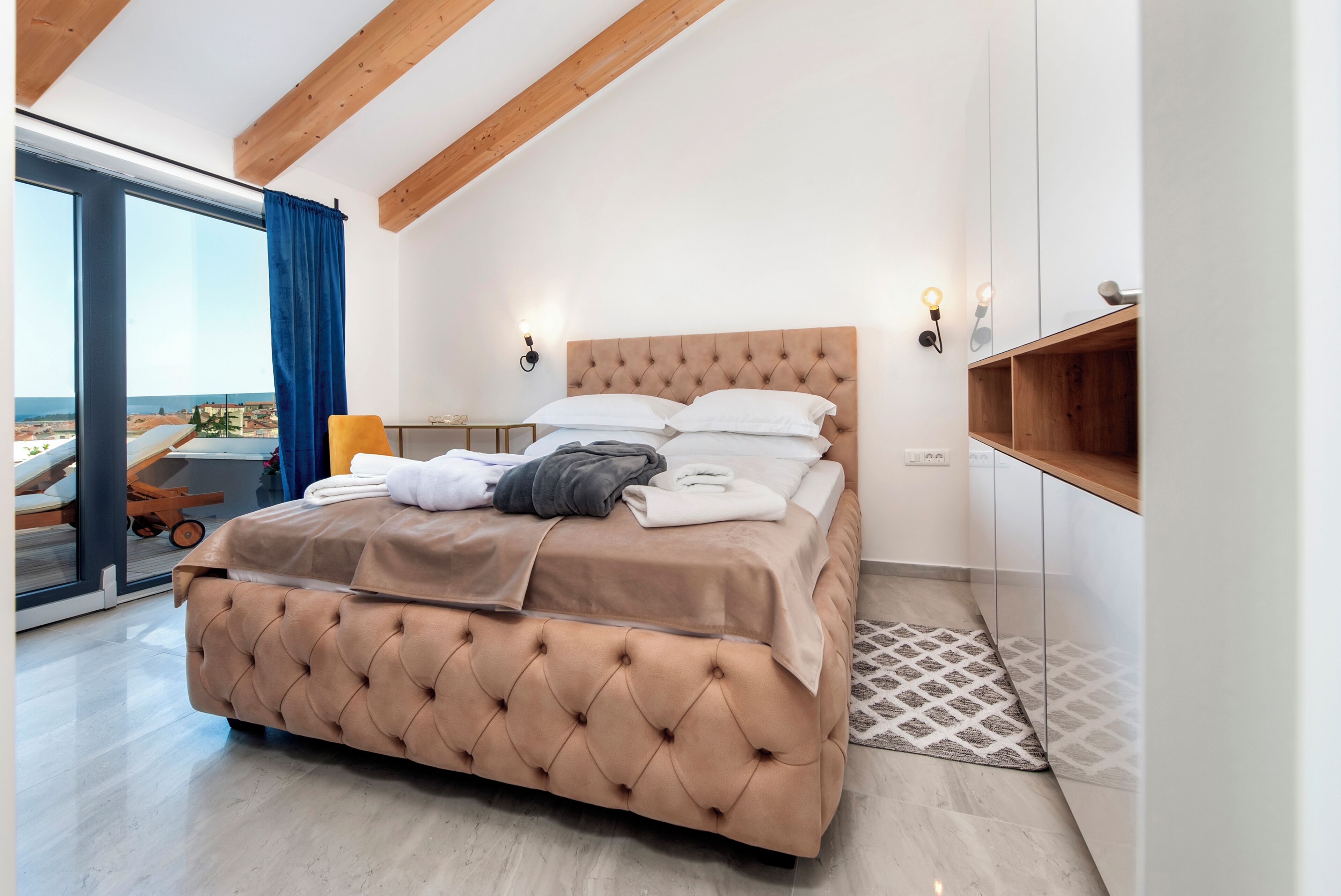 A bedroom with a large bed, a wooden ceiling, a view of the sea, and a small table with a chair.