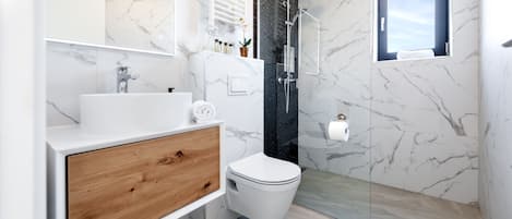 Un baño moderno con pared de mármol, tocador de madera, inodoro blanco, cabina de ducha con mampara de cristal y una ventana pequeña.