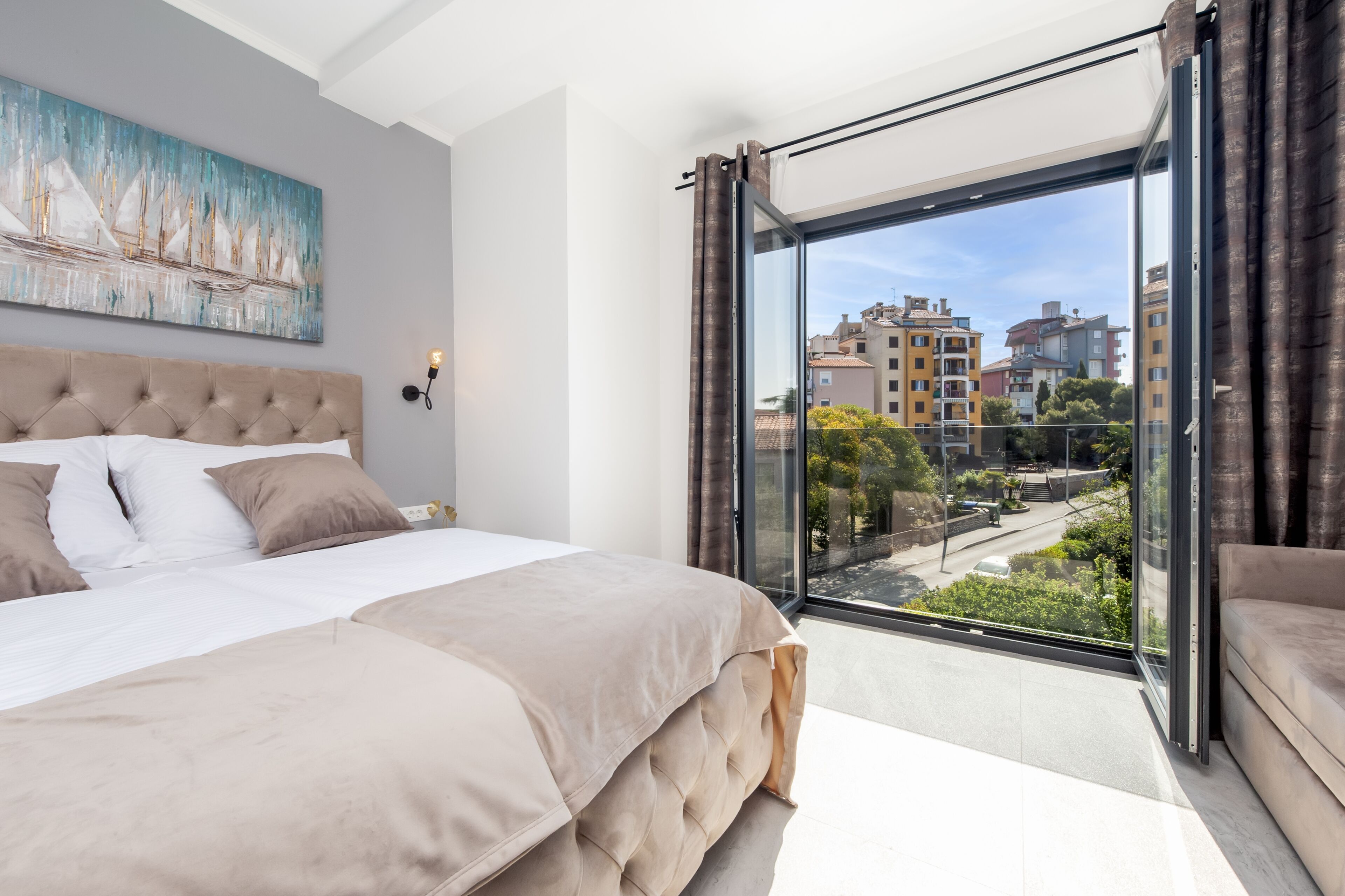 A modern bedroom with a large bed, a city view through floor-to-ceiling windows, and a painting on the wall.