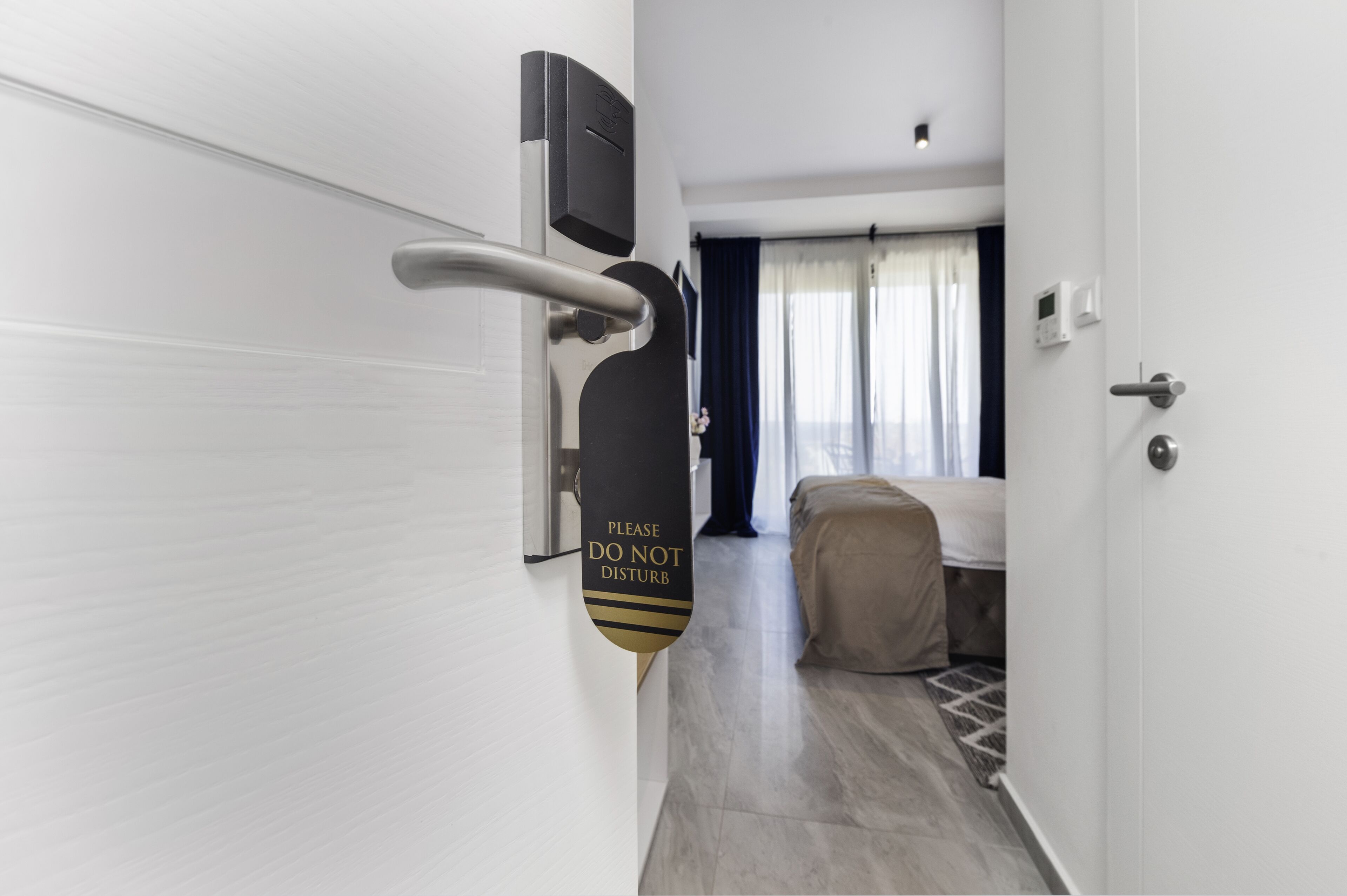 A hotel corridor with a bed, a door, and a 'Please Do Not Disturb' sign.
