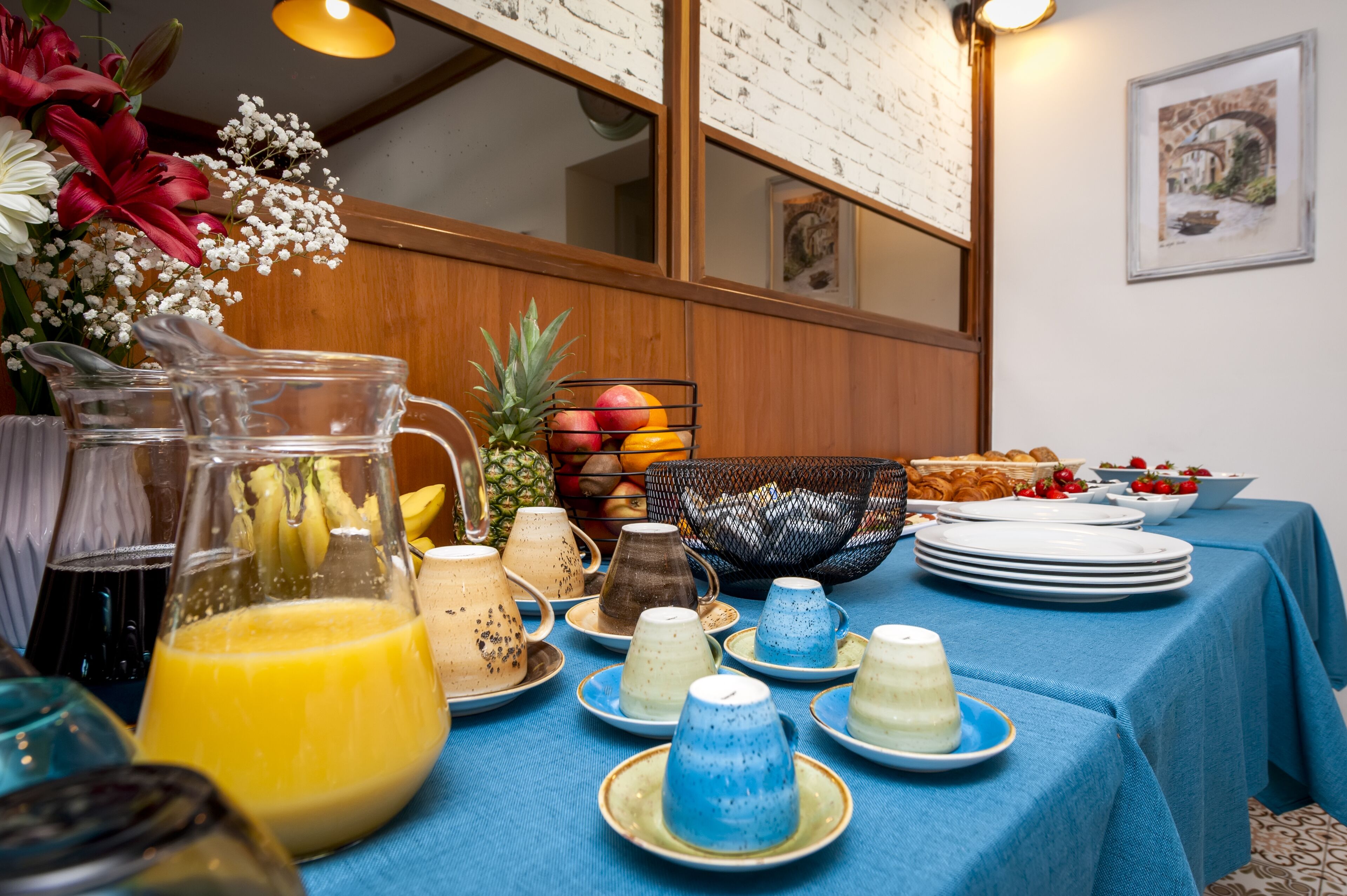Una mesa de desayuno con diversas bebidas y frutas, una cesta de pan y un mantel azul.