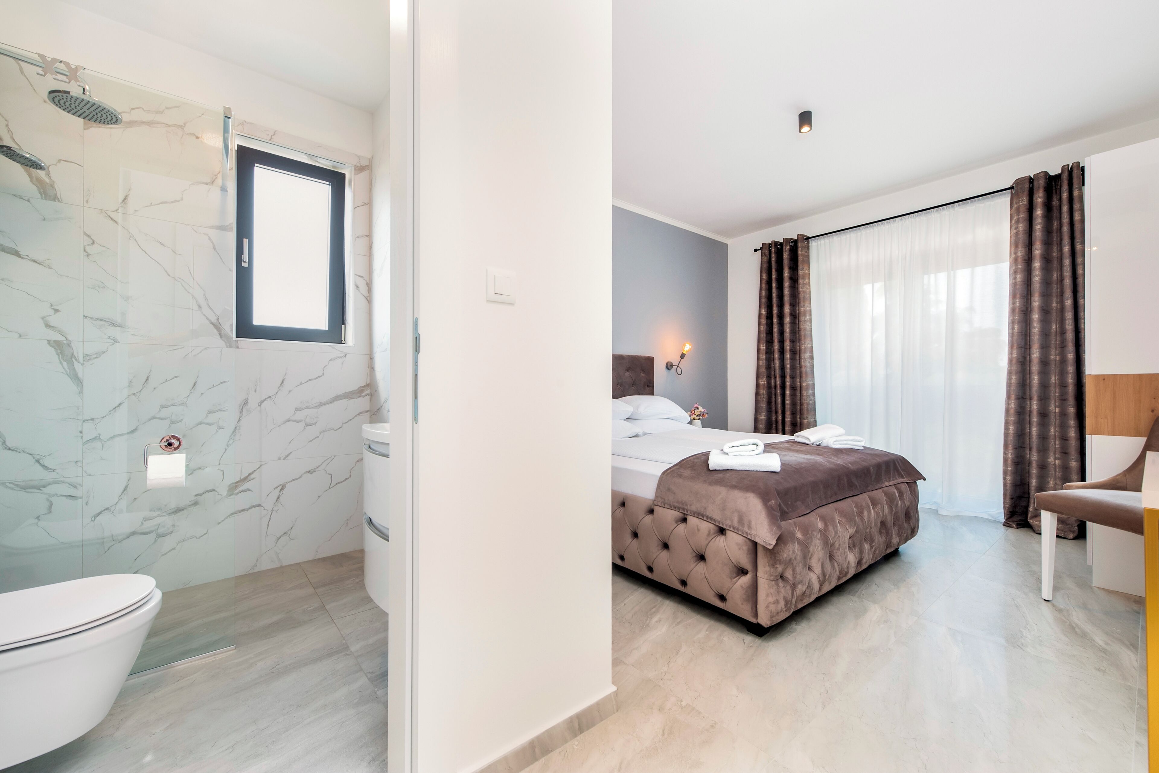 A modern hotel room with a marble bathroom, a bed with a brown headboard, and a large window with curtains.