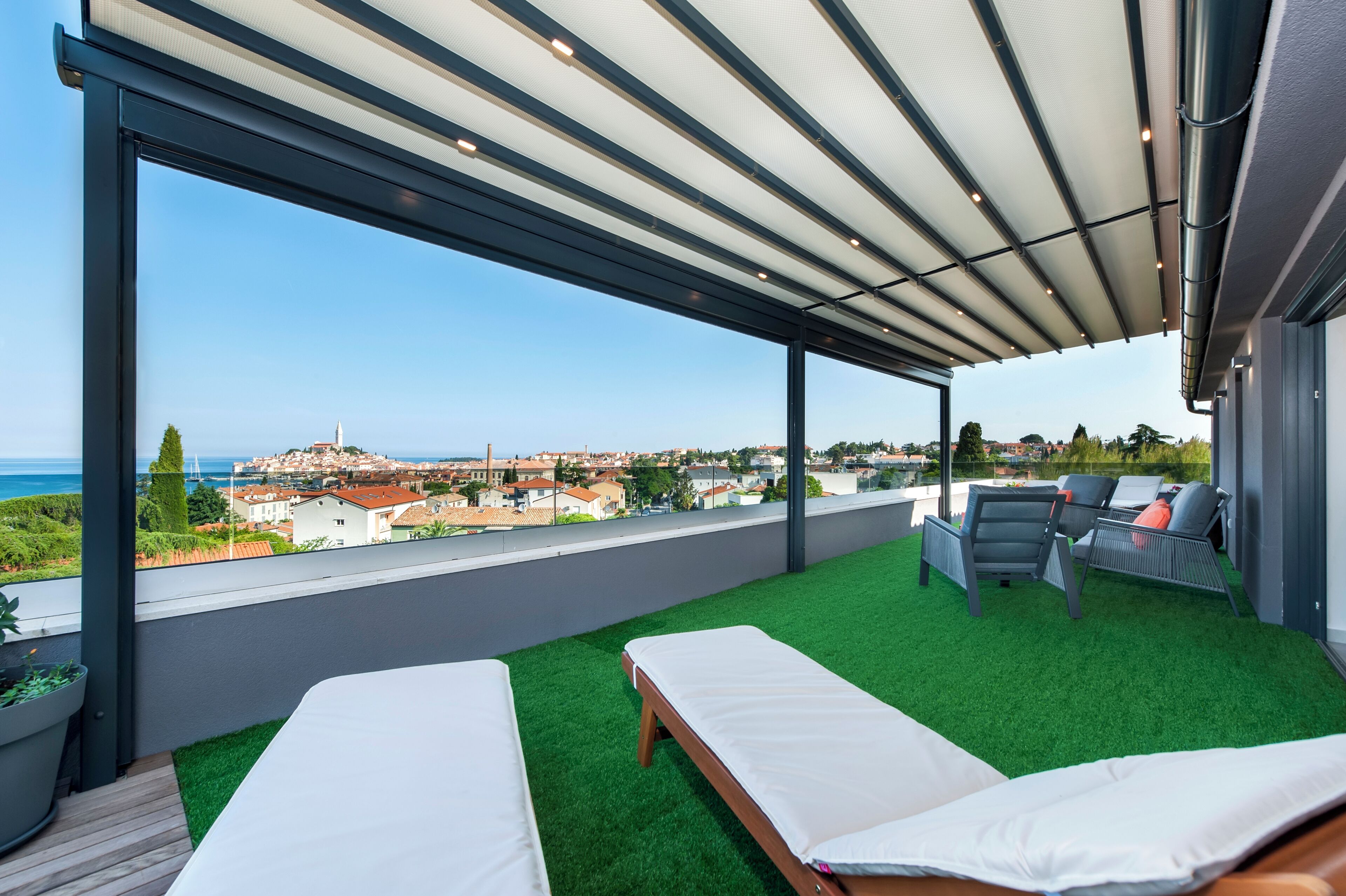 Een modern balkon buiten met twee bedden, een zithoek en uitzicht op zee en de stad.