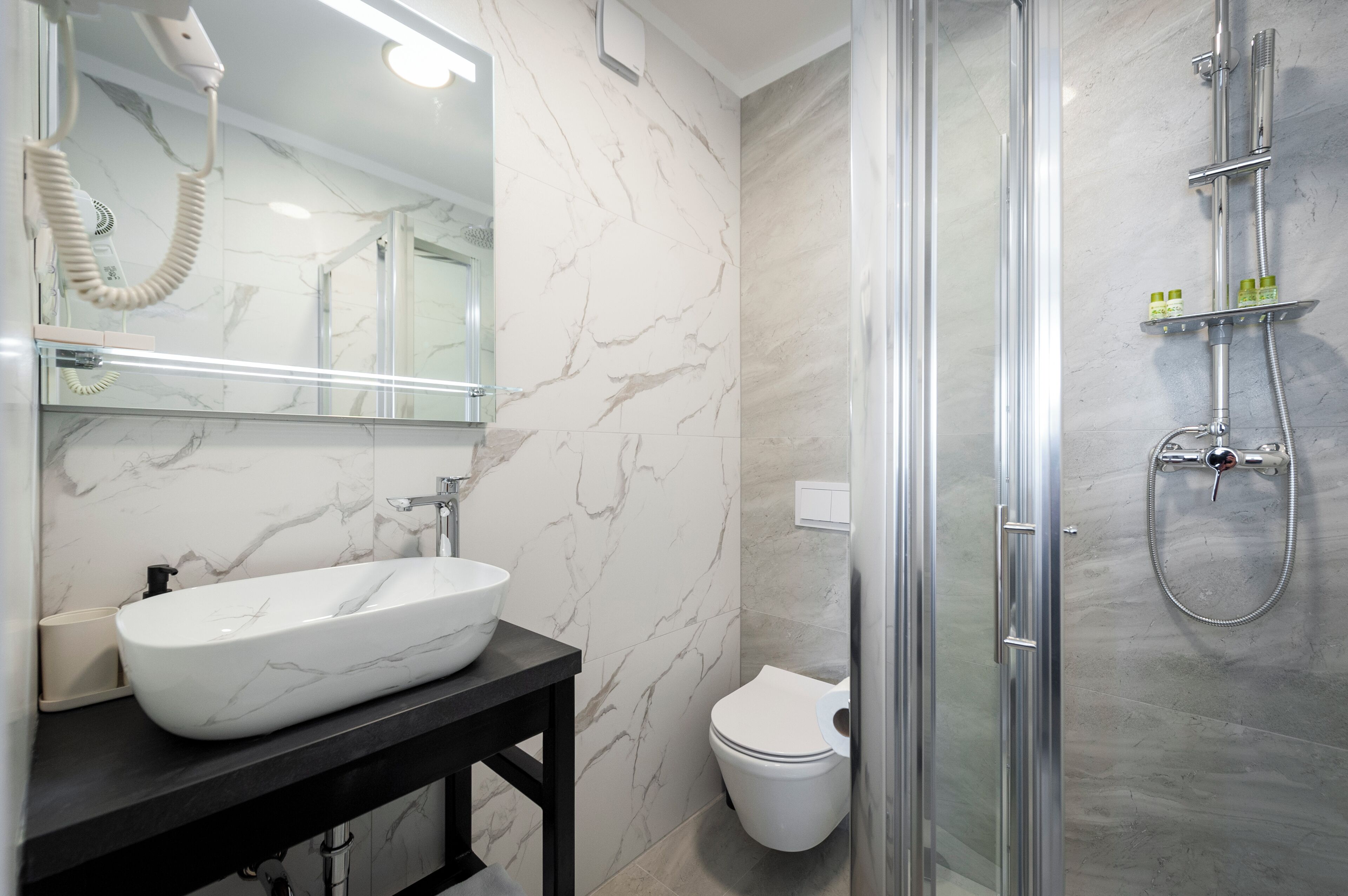 Un baño con lavamanos de mármol blanco, tocador negro, inodoro blanco y zona de ducha con cerramiento de vidrio.