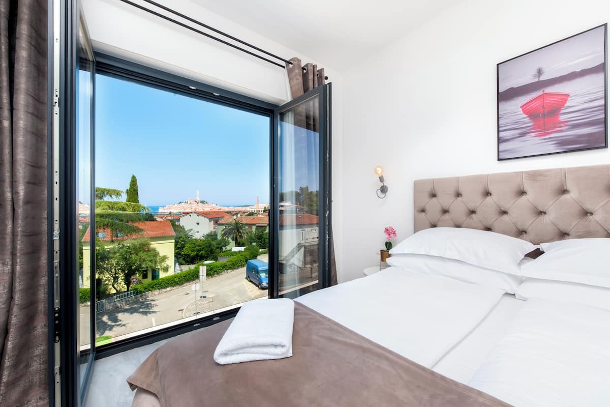 A hotel room with a large window, a bed with white and brown bedding, a tufted headboard, and a framed picture on the wall.