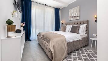 Une chambre à coucher moderne avec un grand lit, une table de chevet, un téléviseur fixé au mur et une fenêtre avec des rideaux.