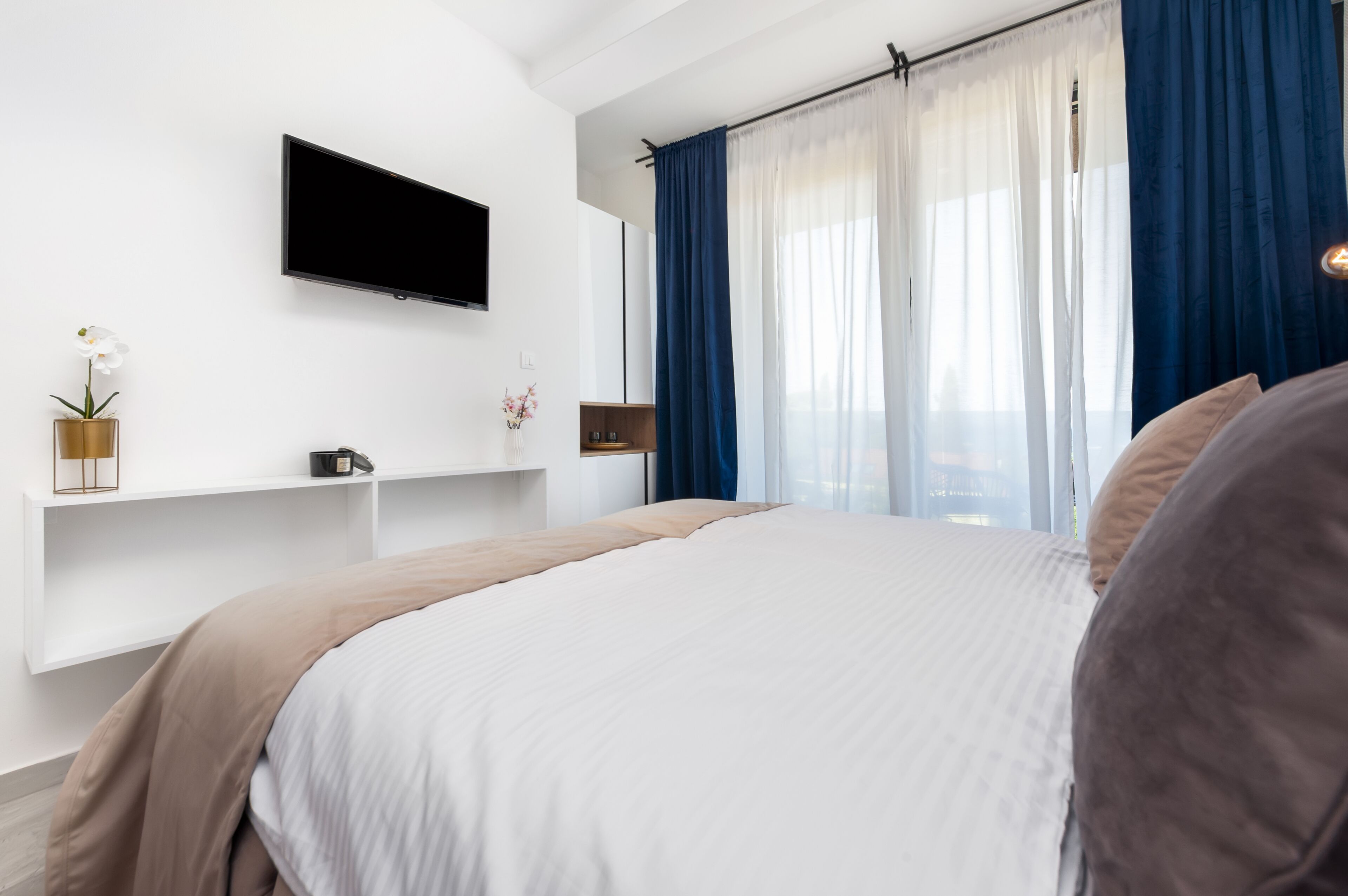 Un dormitorio moderno con una cama grande, un televisor montado y una ventana con cortinas azules.