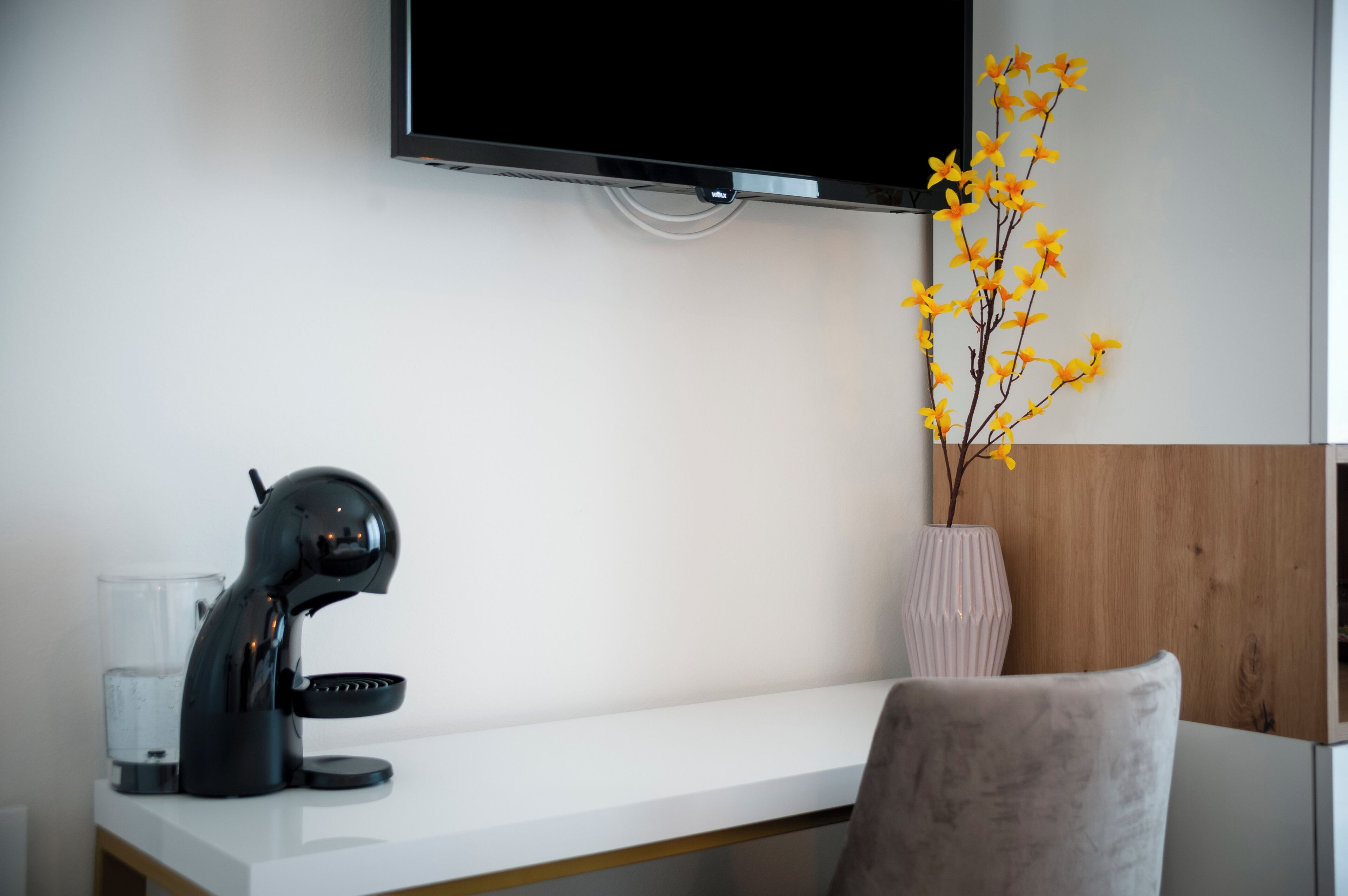 Ein modernes Hotelzimmer mit weißem Schreibtisch, einer schwarzen Schreibtischlampe, einem klaren Glas, einer rosa Vase mit gelben Blumen und einem an der Wand montierten Fernseher.