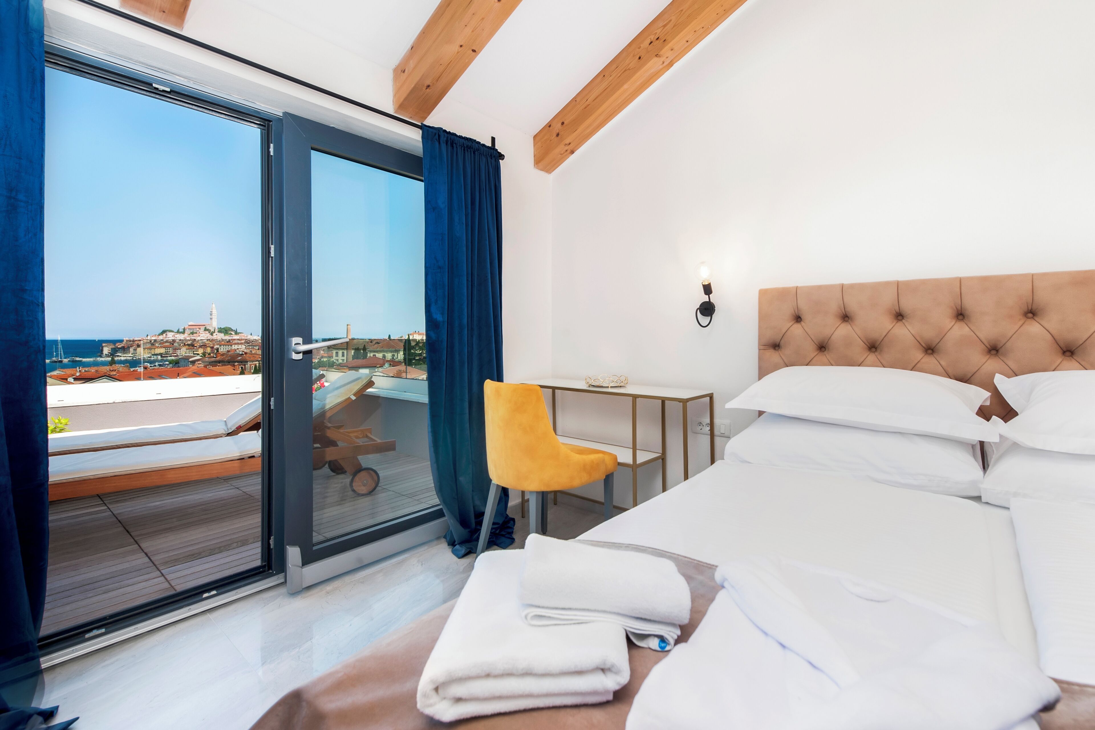 A hotel room with a bed, a desk, a chair, and a view of a coastal town through a glass door.