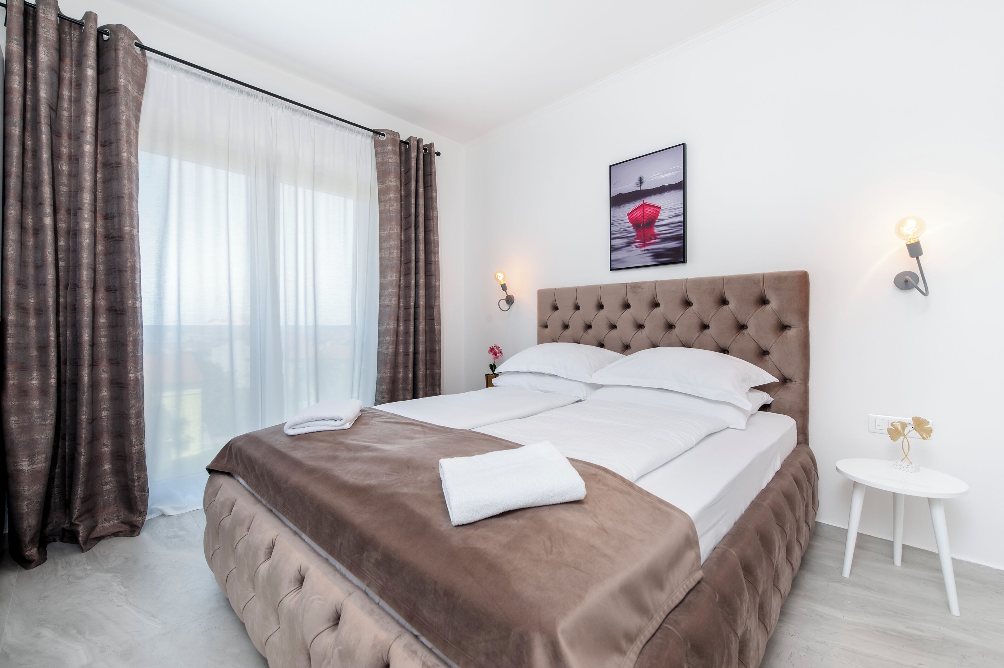 Una cama bien hecha con un cabecero marrón, sábanas blancas y una toalla blanca sobre la cama. Junto a la cama hay una mesita pequeña con un adorno encima. Una fotografía cuelga de la pared sobre la cama.