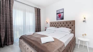 A neatly made bed with a brown headboard, white linens, and a white towel on the bed. There is a small white table next to the bed with a decorative item on it. A picture hangs on the wall above the bed.