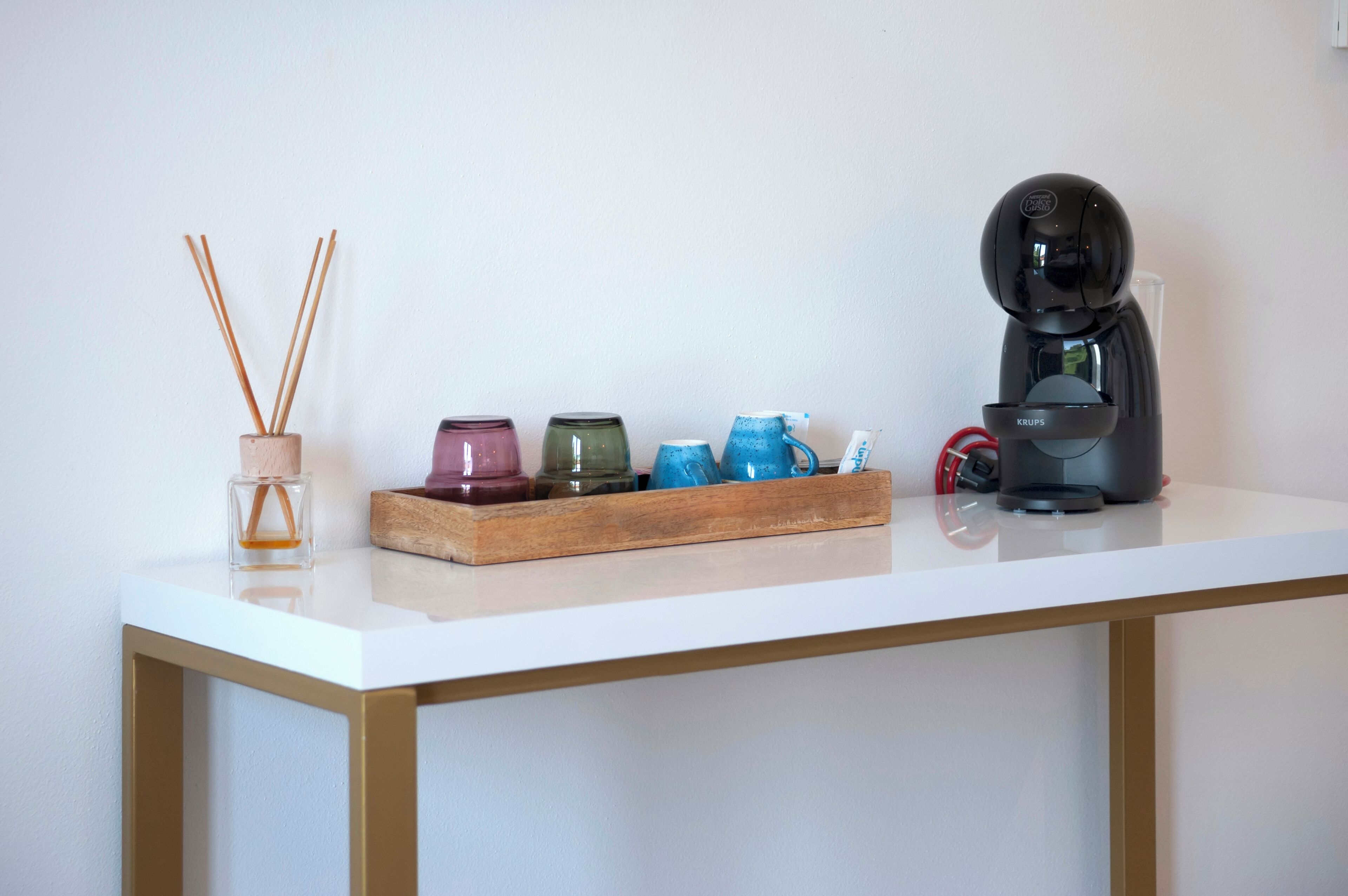 Un tavolo bianco con un vassoio di legno che contiene cinque contenitori di vetro colorati, una macchina da caffè nera e un piccolo barattolo di vetro con bastoncini di canna.