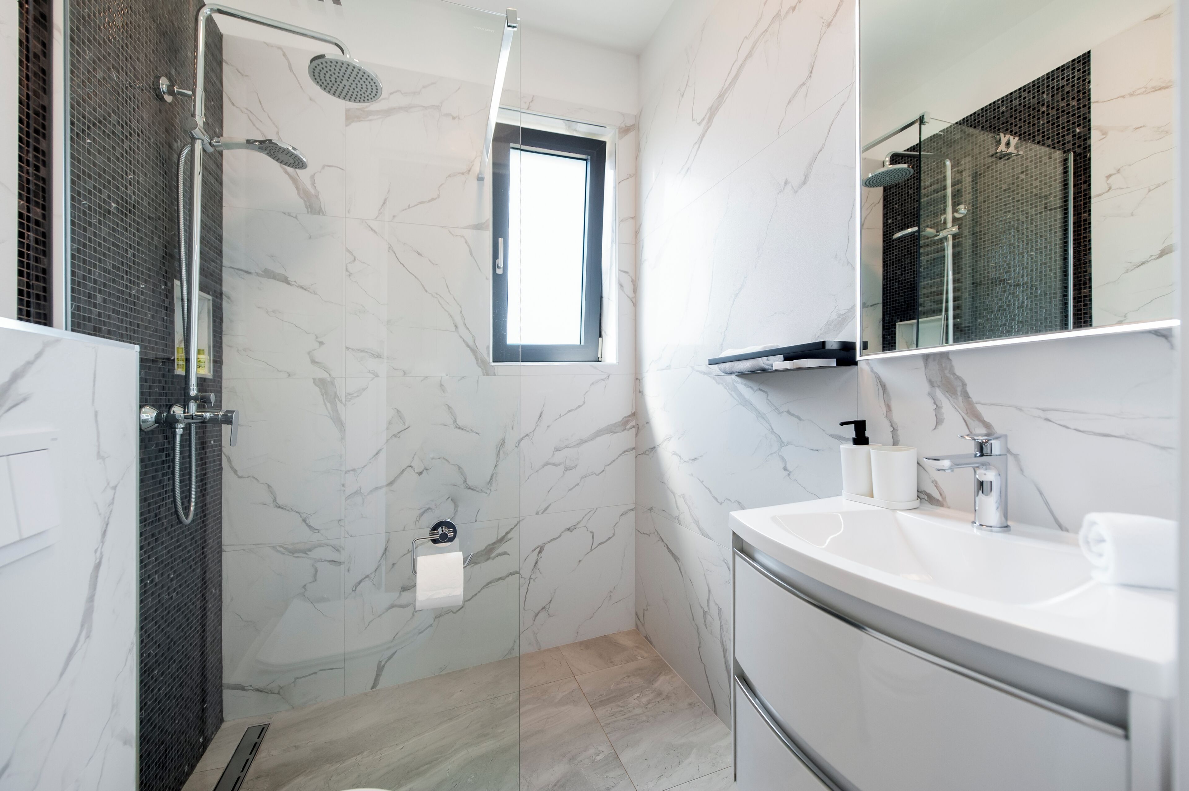 Un baño moderno con cabina de ducha de vidrio, pared de mármol, lavabo con espejo y ventana que proporciona luz natural.