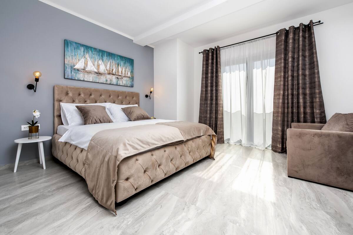 A bedroom with a large bed, a side table, a potted plant, and a painting on the wall.