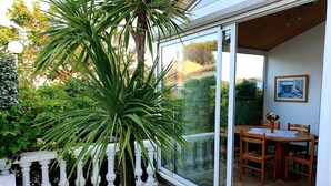Eine Terrasse mit Glastür zu einem Essbereich im Freien, umgeben von Grünpflanzen und einem weißen Geländer.