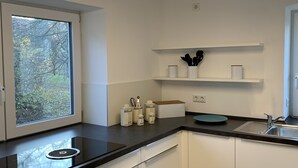 Moderni keittiö, jossa on valkoiset kaapit, musta työtaso, ikkuna, josta avautuu näkymä puille, sekä sisäänrakennettu hylly keittiövälineille.