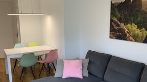 Moderni olohuone, jossa on harmaa kulmasohva, värikkäillä tuoleilla varustettu ruokapöytä ja suuri maisemamaalaus seinällä.